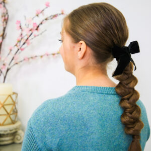 Side view of girl standing indoors with a blue shirt modeling "Wrapped Braid" hairstyle with a black bow