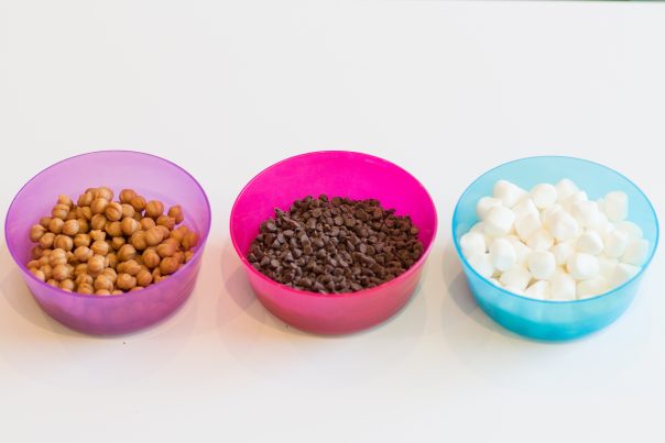(Left to right) Purple bowl filled with caramel, pink bowl filled with chocolate chips, and blue bowl filled with marshmallows