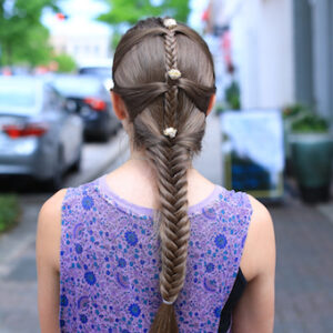 Back view of girl standing outside with a purple shirt modeling "Fishtail Mermaid Braid" hairstyle with flower accessories.