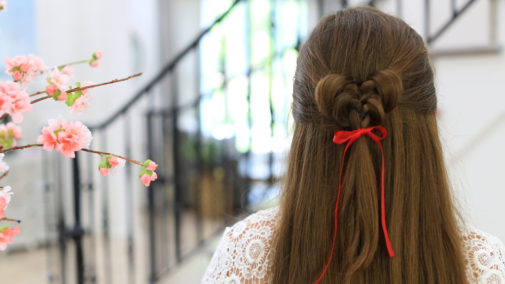 Close up back view of girl standing indoors wearing a red ribbon in her hair modeling the "Butterfly Tieback" hairstyle