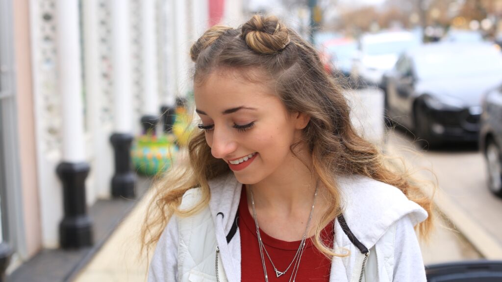 Side view of smiling girl looking down, wearing a red shirt with a white jacket modeling "Double Bun Half Up" hairstyle