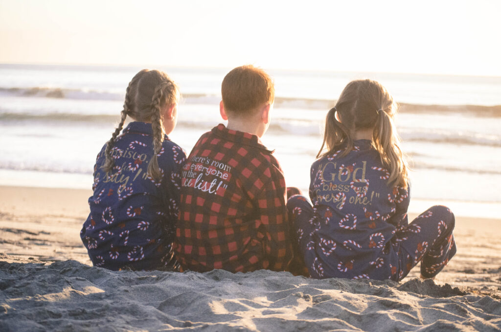 Two girls and one boy in the middle sitting on the beach in personalized Christmas pajamas
