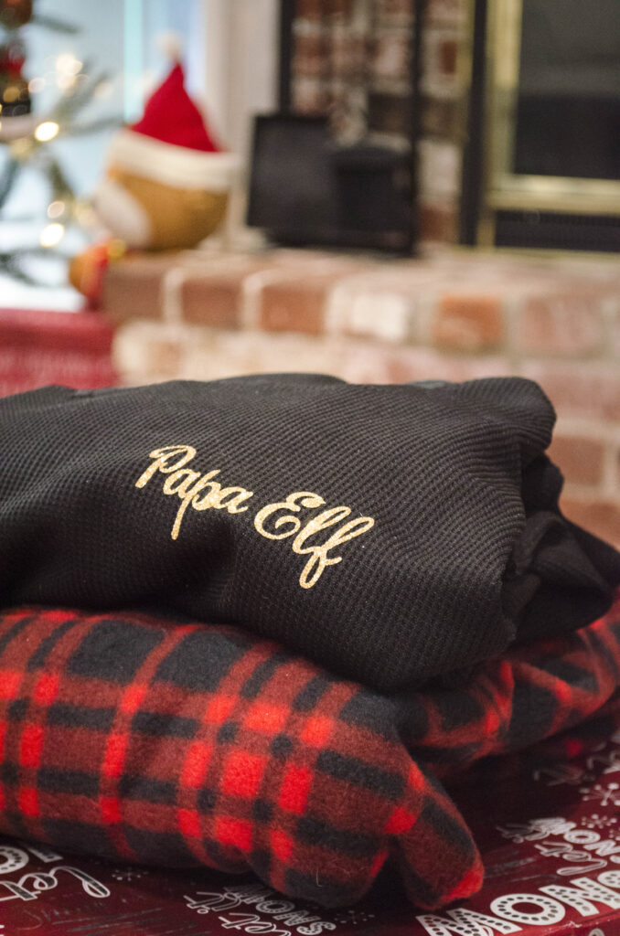 Black 'Papa Elf' shirt stack on plaid blanket on top of Christmas presents