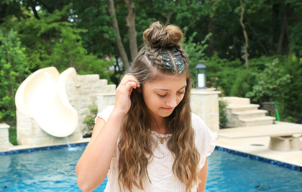 Girl standing by the pool with glitter in her hair modeling Double Dutchbacks into a Messy Bun hairstyle