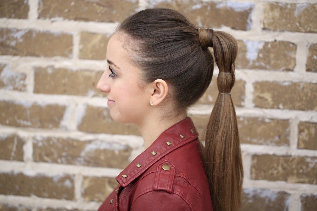 Profile of girl standing outside in front of a brick wall wearing a red jacket modeling "Heart Ponytail" hairstyle