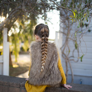 Back view of girl wearing faux fur vest over yellow dress outside modeling the "Dutch Pull-Thru Combo Braid" hairstyle