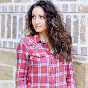 Woman with curling hair wearing a red plaid shirt standing outside in front of a brick background