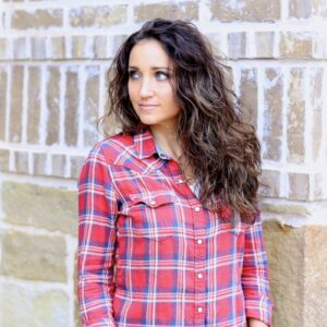 Woman with curly hair wearing a red plaid shirt standing outside in front of a brick background