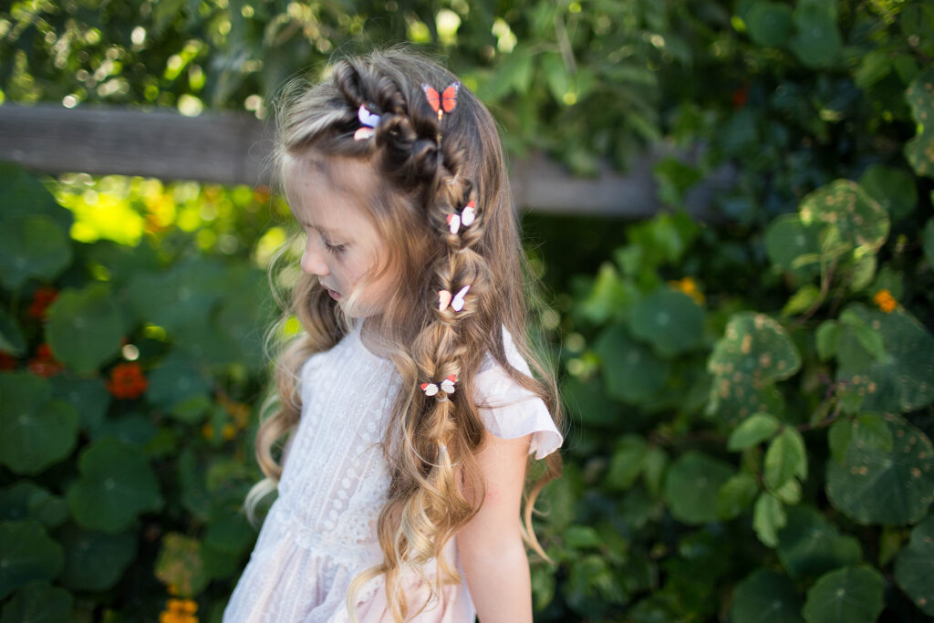 Side view of a young girl with hair standing outside modeling "Flip Faux Fishtail" hairstyle with butterfly accessories