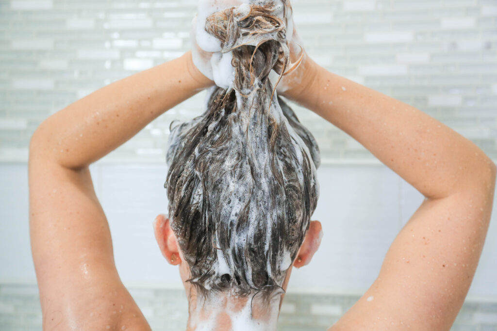 Back view of a woman holding her hair while washing her hair full of suds with both hands.